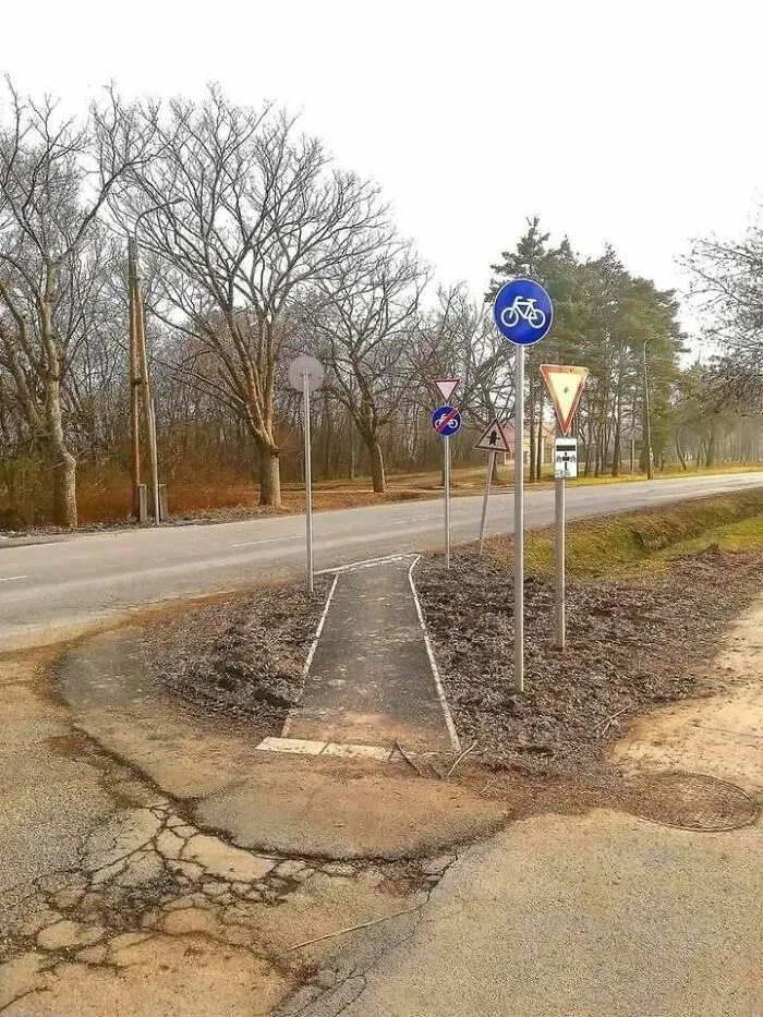 Narrow bike path surrounded by multiple confusing traffic signs in a rural Eastern Europe setting.