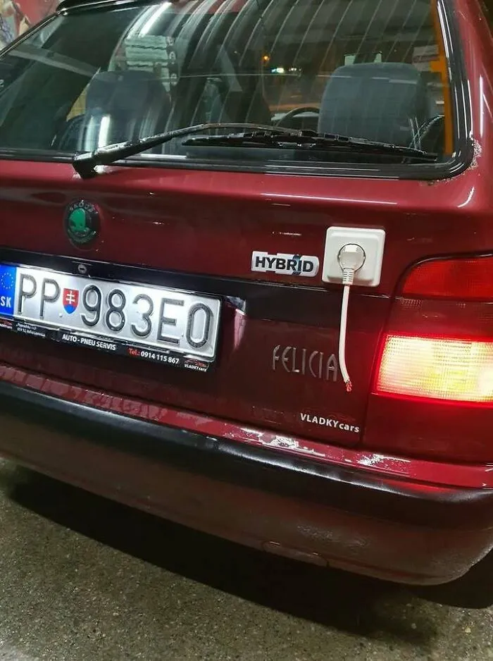 Red car with cracked bumper and a fake hybrid badge with an electrical plug socket attached, illustrating Eastern Europe humor.