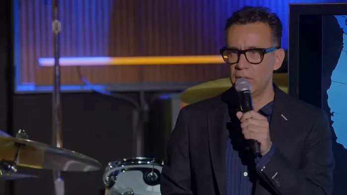 Man in glasses speaking into a microphone on stage, with drums and a monitor visible, discussing celebrities’ closet skeletons.