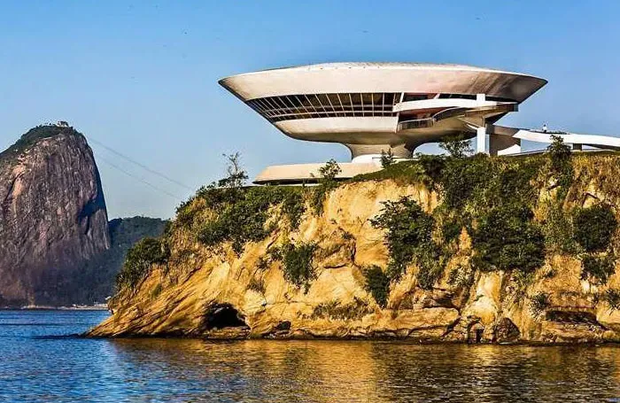 Futuristic bread-shaped bread factory building perched on a rocky coastline with clear blue sky and water.
