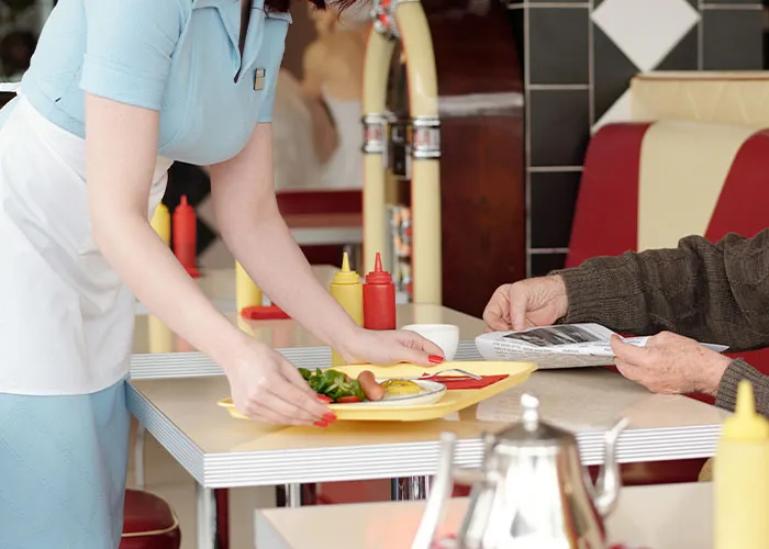Waitress serving a meal with ketchup and mustard bottles on the table in a chaotic diner scene.