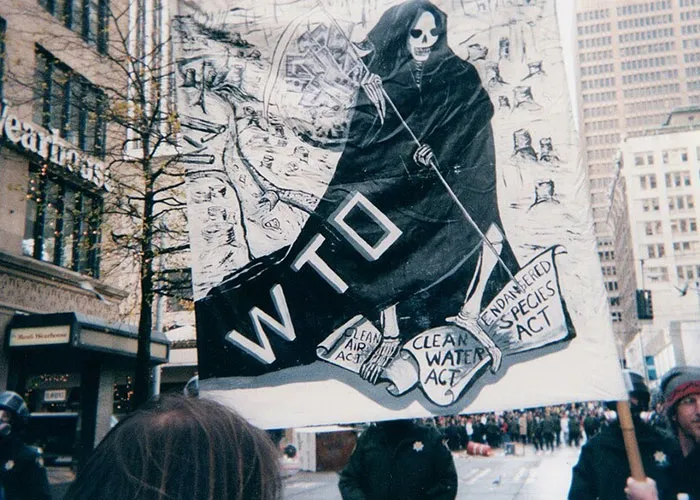 Protesters holding chaotic scenes banner depicting grim reaper with WTO and environmental acts in a city street demonstration.