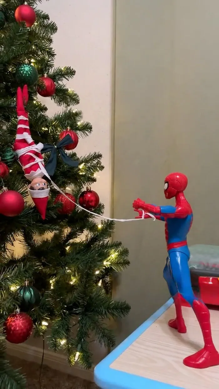 Elf on the Shelf tied to Christmas tree by Spider-Man toy creating a mischievous holiday scene with decorations and lights.