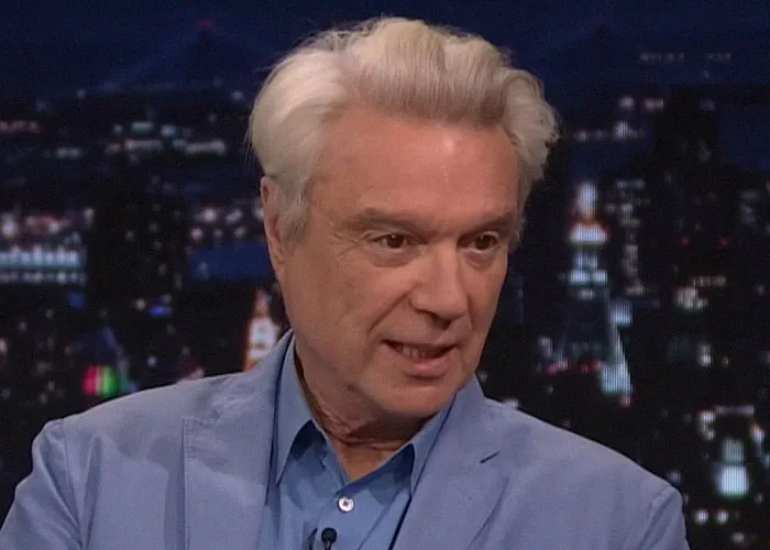Man with white hair wearing a blue blazer speaking about meeting famous people and sharing celebrity experiences on a nighttime show.