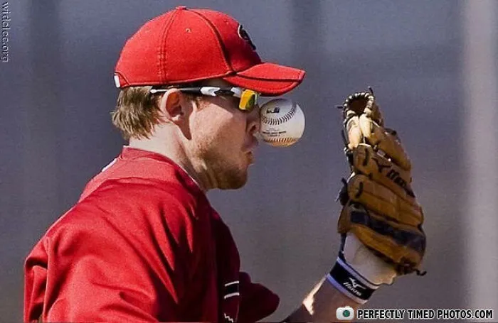 Baseball player in red cap and glove caught in a perfectly timed photo as the ball is about to hit his face.