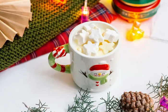 Festive holiday mug with marshmallows surrounded by Christmas decorations and cozy knitwear on a white table.