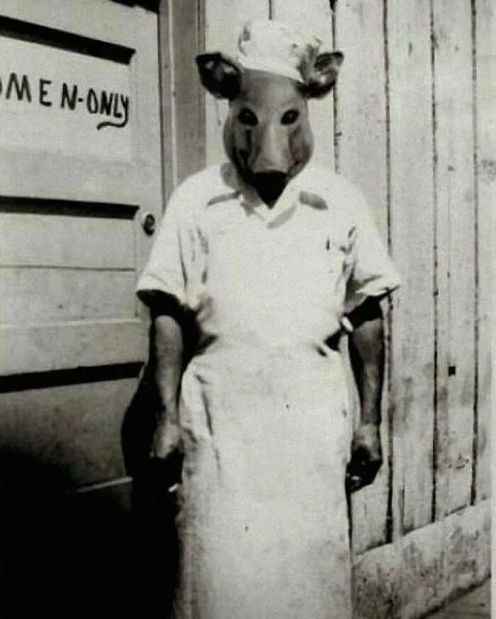 Black and white vintage photo of a person wearing a creepy animal mask and chef uniform standing by a women-only door.