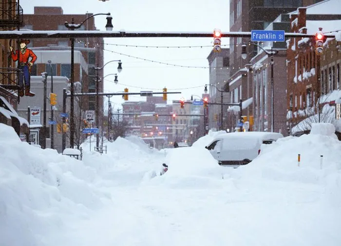 Urban street covered in deep snow during a heavy snowstorm hitting the U.S. with blocked cars and traffic lights visible.