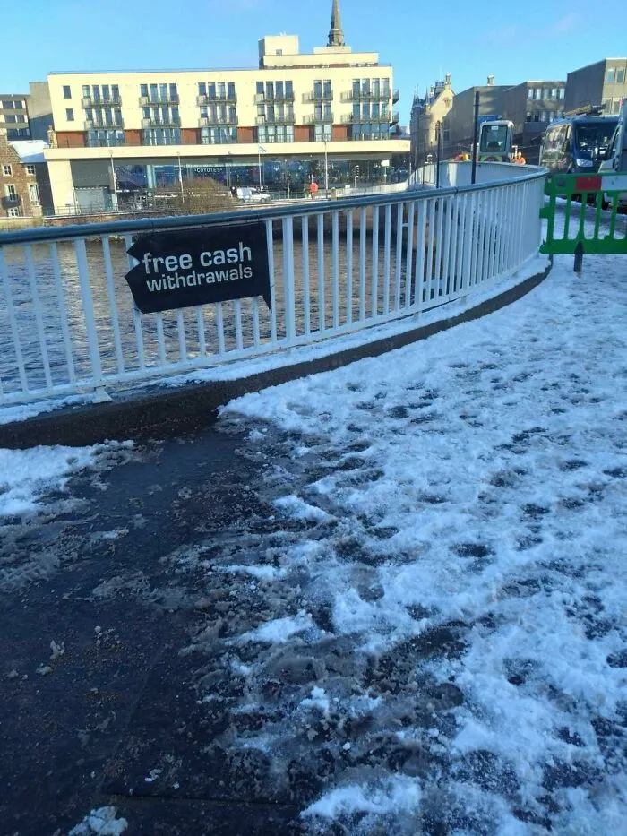 Black sign reading free cash withdrawals attached to railings on a snowy path near a river in a tourist area.