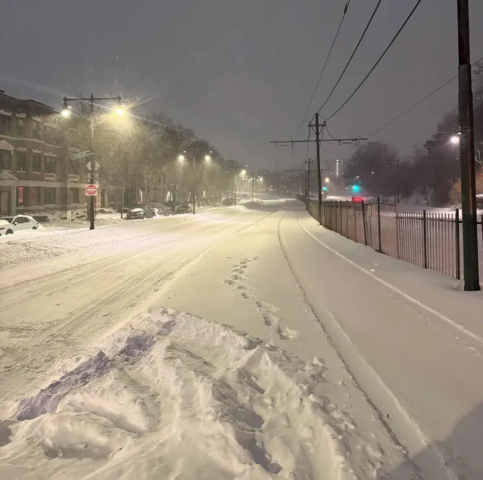 Snowstorm hitting a U.S. city street at night with snow-covered road, sidewalk, and streetlights glowing in the snowfall.