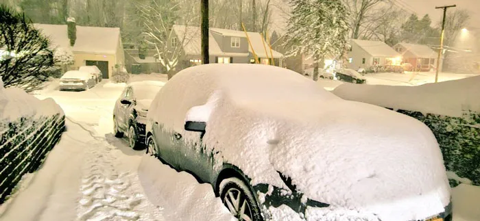 Car covered in thick snow during a heavy snowstorm in a residential U.S. neighborhood at night.
