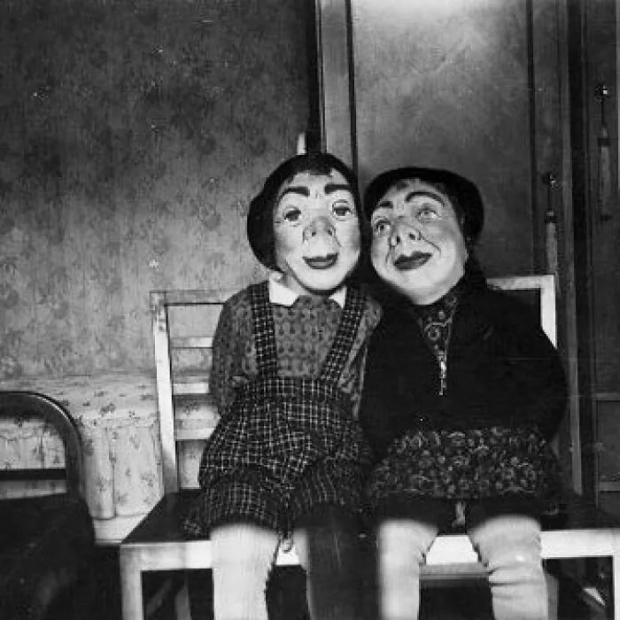 Two old black and white creepy dolls with painted faces sitting side by side on chairs in a dimly lit room.