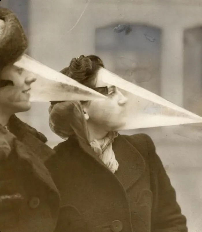 Two women in old black and white photo wearing long, pointed odd and creepy face masks outdoors.