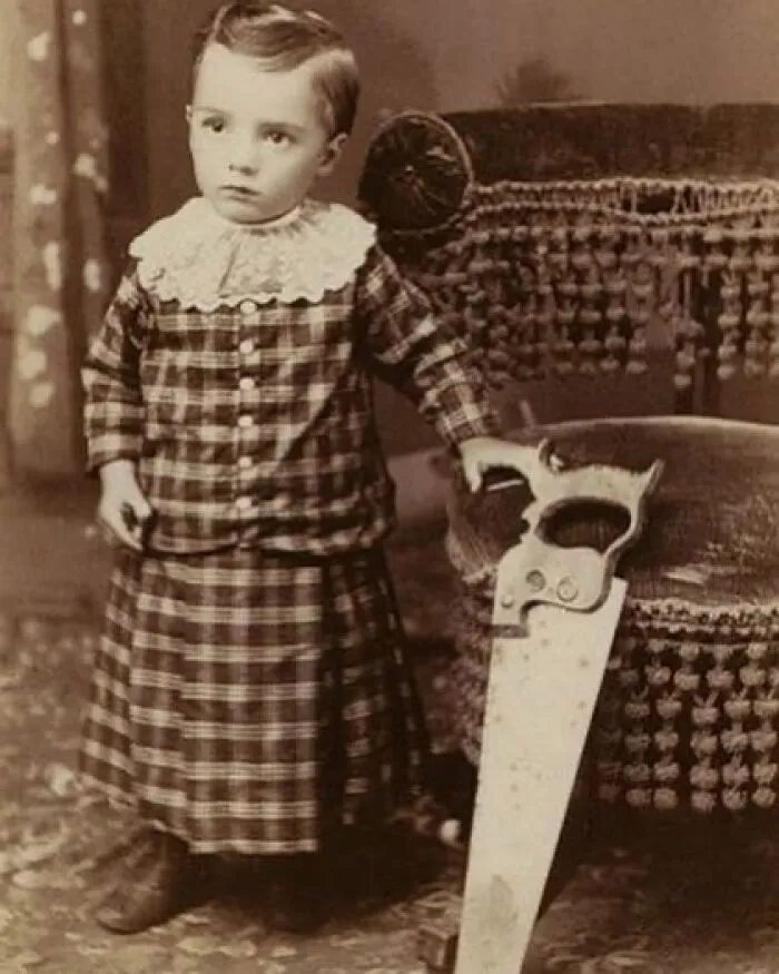 Vintage black and white creepy photo of a young child in old-fashioned clothing holding a large saw odd black and white pic.