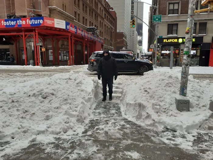 Person bundled in winter clothing walking on a cleared path amid heavy snowstorm accumulation in a U.S. city street.