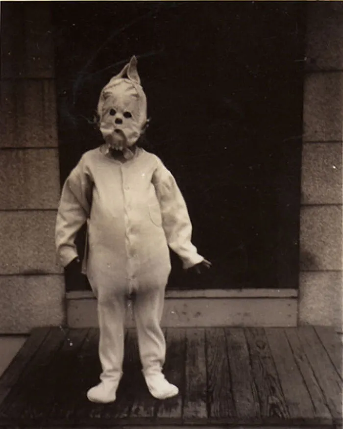 Child in an old black and white creepy costume standing on a wooden porch, evoking odd and eerie feelings.
