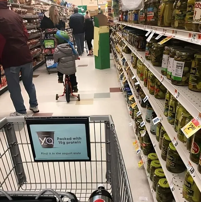 Shopping cart view down grocery aisle with child riding tricycle past shoppers and pickle shelves — next-level jerks moment