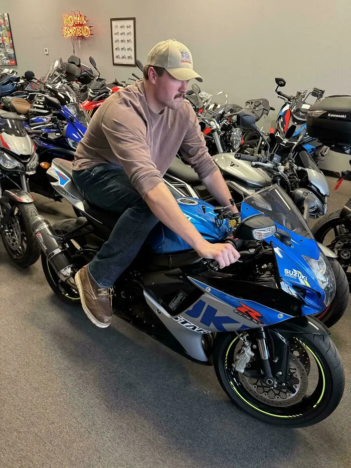 Tall man struggles to fit comfortably on a blue Suzuki motorcycle in a showroom filled with bikes.