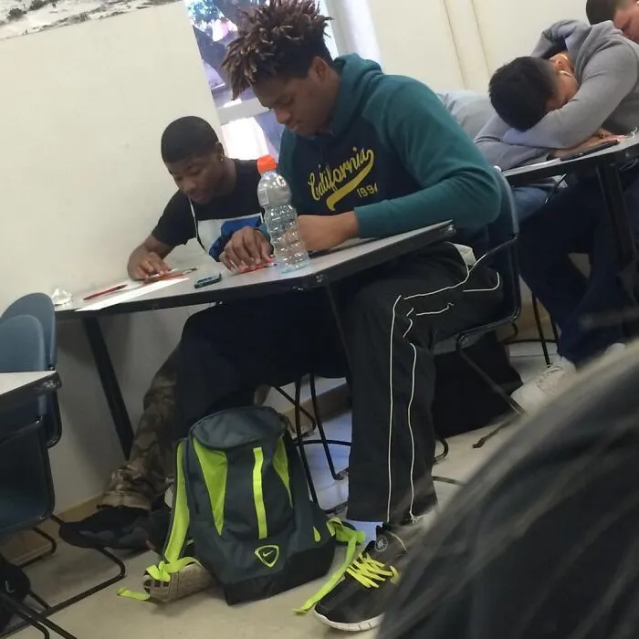 Two tall students sitting at desks in a classroom, one wearing a blue hoodie and struggling with legroom.
