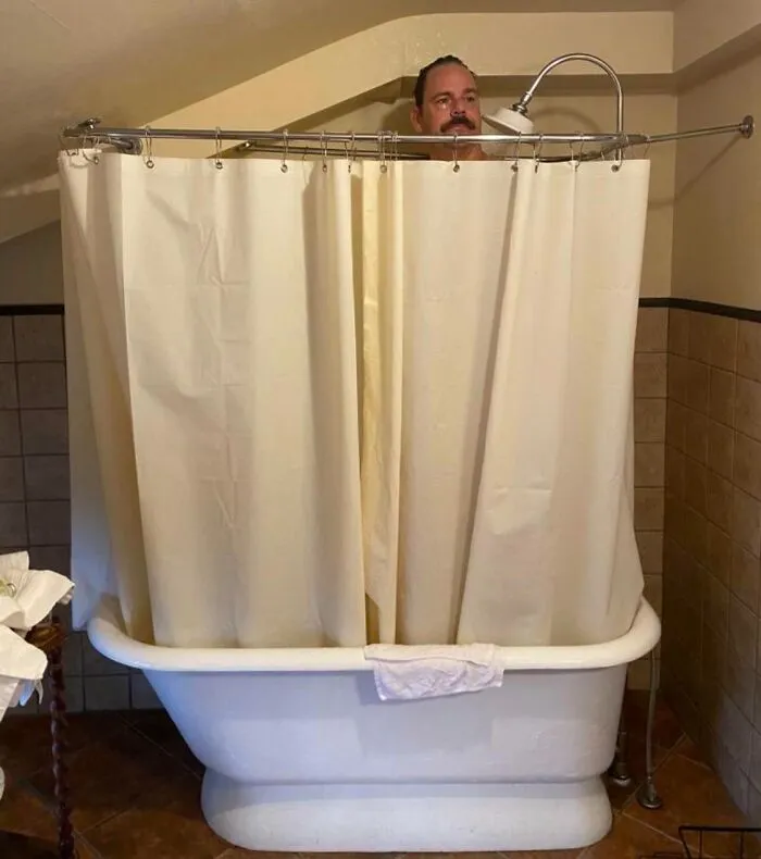 Man in a bathtub with shower curtain barely covering him, illustrating common struggles tall people face in bathrooms.
