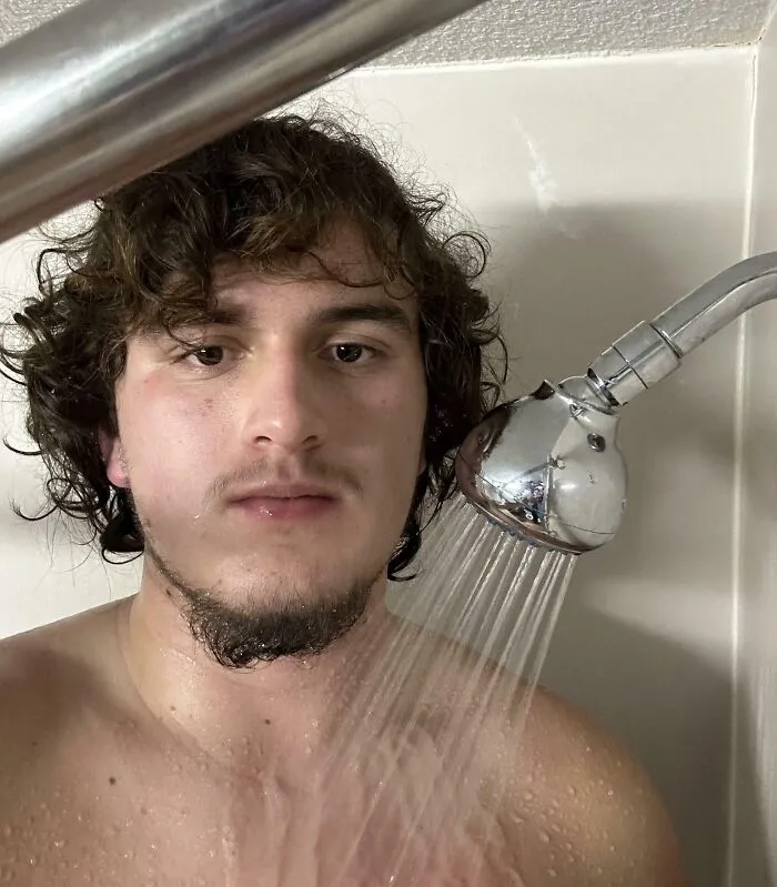 Young man struggling with short showerhead in bathroom, illustrating common struggles only tall people face.