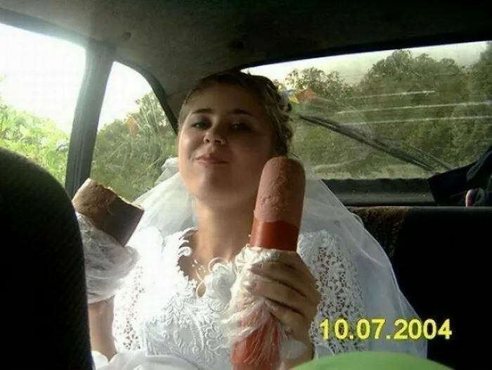 Bride in wedding dress holding two large sausages inside a car, an awkward wedding photo that is both funny and cringe-worthy.