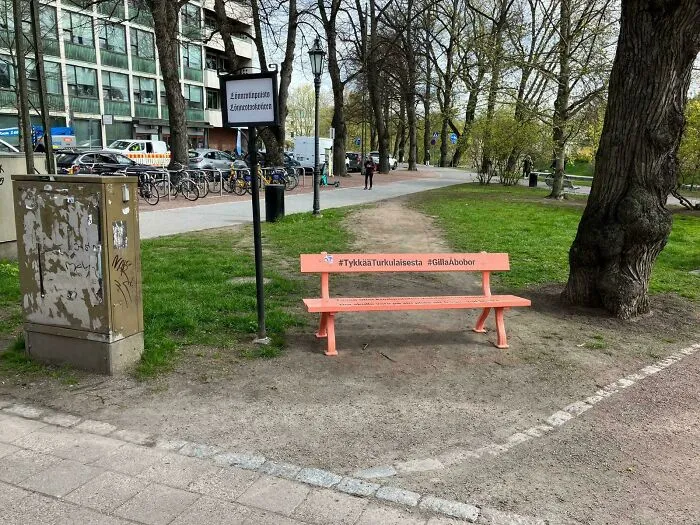 Desire path worn in grass alongside paved walkway near an orange bench in an urban city park area