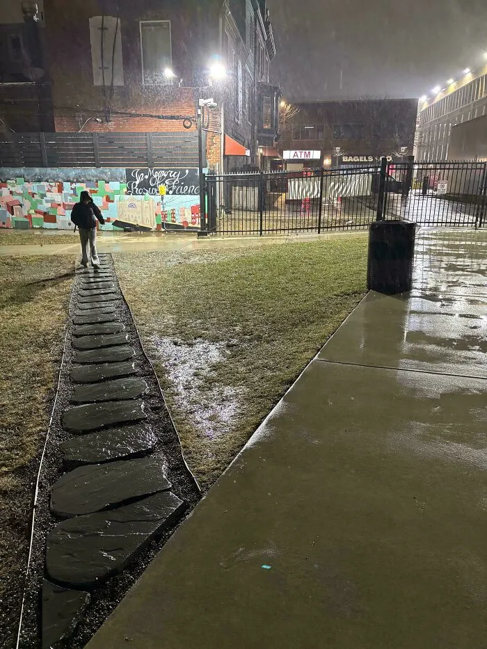 Person walking on a wet desire path made of stones next to a sidewalk, illustrating how cities plan versus actual pedestrian routes.