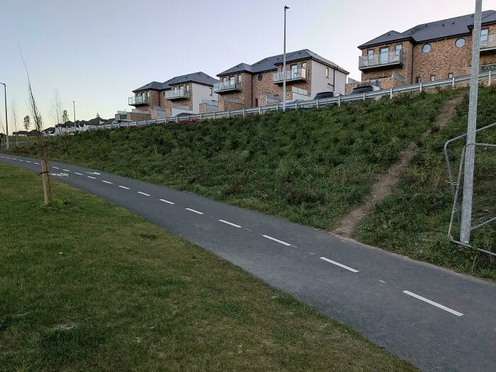 Desire path cutting diagonally across grassy slope beside paved city walking path near residential houses.