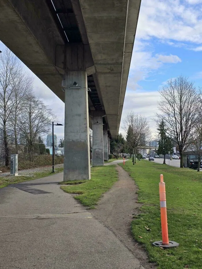 Concrete urban overpass with paved sidewalk and visible desire path worn into grass alongside it.