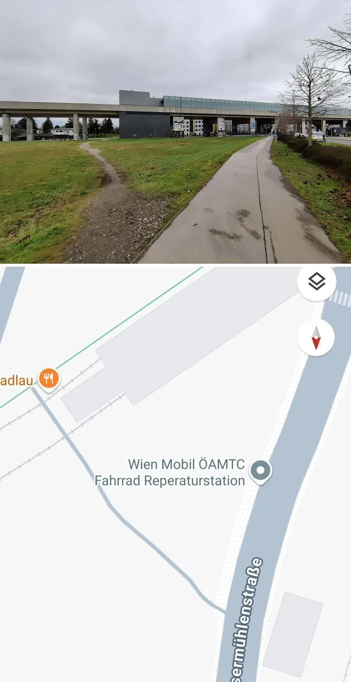 Desire path cutting across grass beside paved walkway near urban area and railway infrastructure in city.