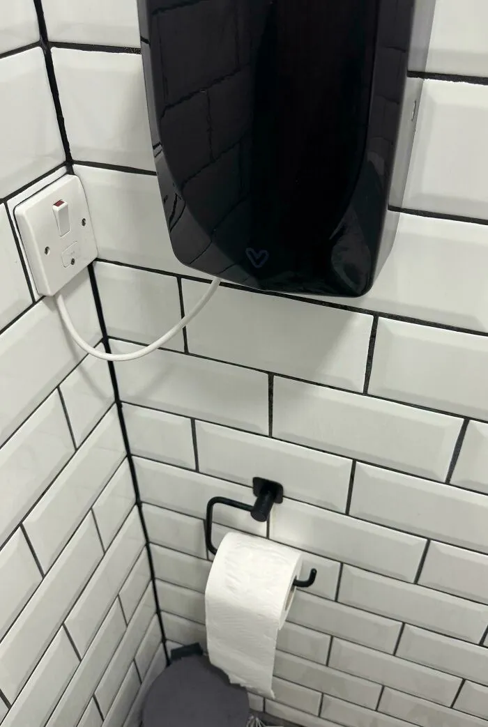 Dispenser negro instalado sobre el papel higiénico en baño con azulejos blancos, error de diseño y mala ingeniería visible.