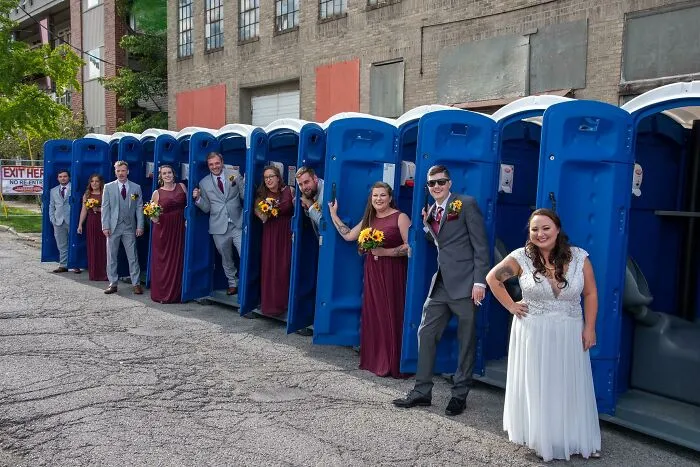 Wedding party in formal attire posing inside and beside a row of portable toilets in an awkward wedding photo.