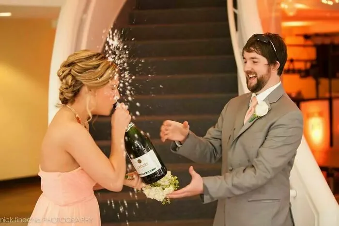 Novia bebiendo champán mientras lo abre y salpica al novio en una foto de boda divertida y fuera de lo común.