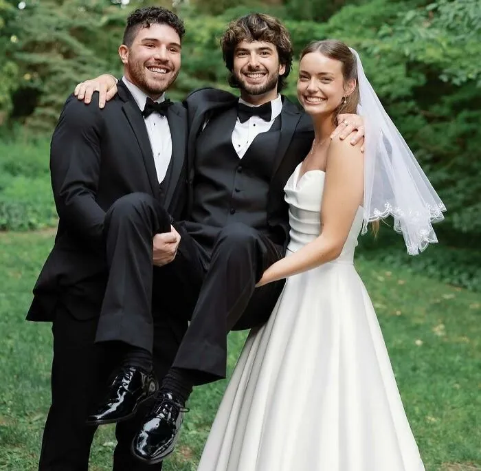 Bride in white gown and veil and two groomsmen lifting another in a tuxedo in awkward wedding photos outdoors.