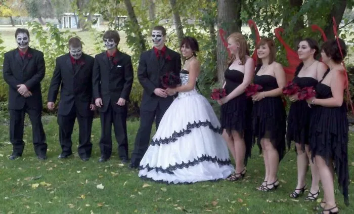 Bride and groom with groomsmen in skull makeup and bridesmaids in black dresses with red devil horns at an awkward wedding photo.