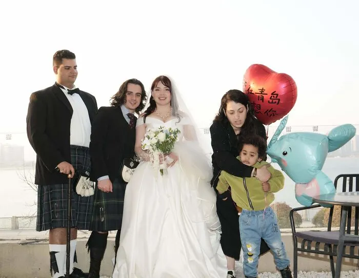 Bride and groom with groomsmen in kilts posing awkwardly next to a woman and child holding a heart-shaped balloon.