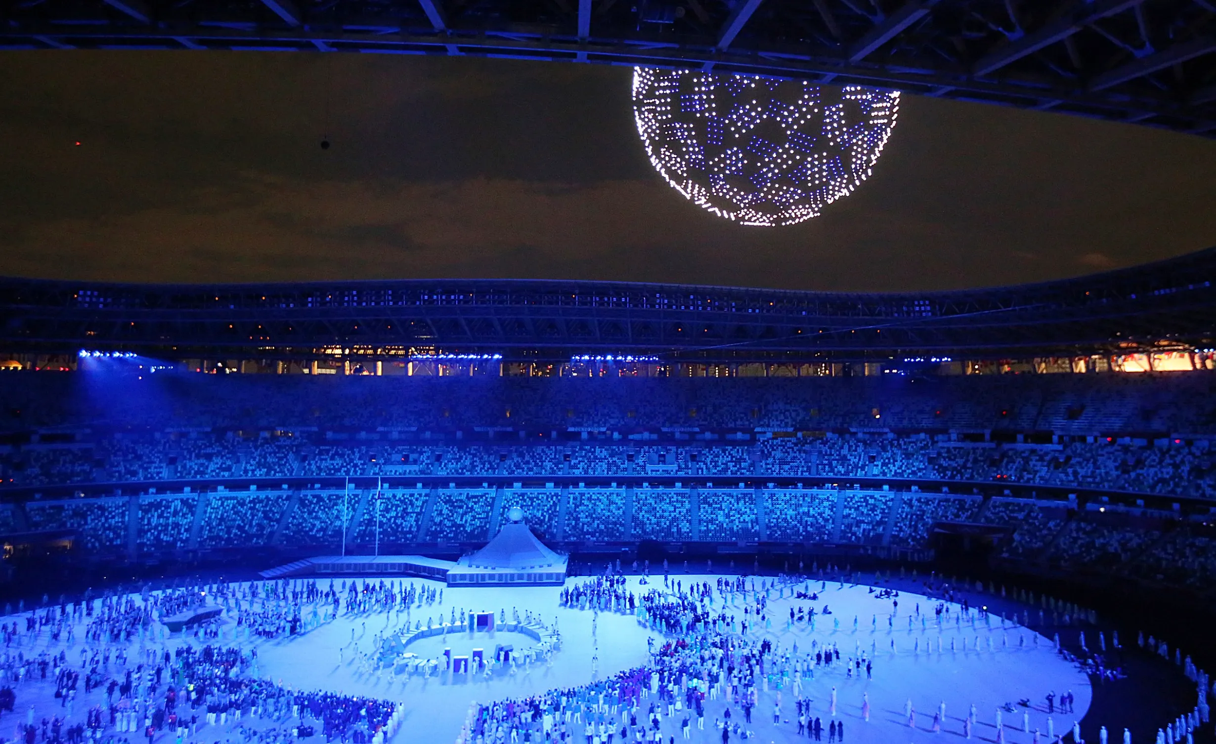 Olympic stadium illuminated in blue with large crowd and drone light display during Summer Olympics event.