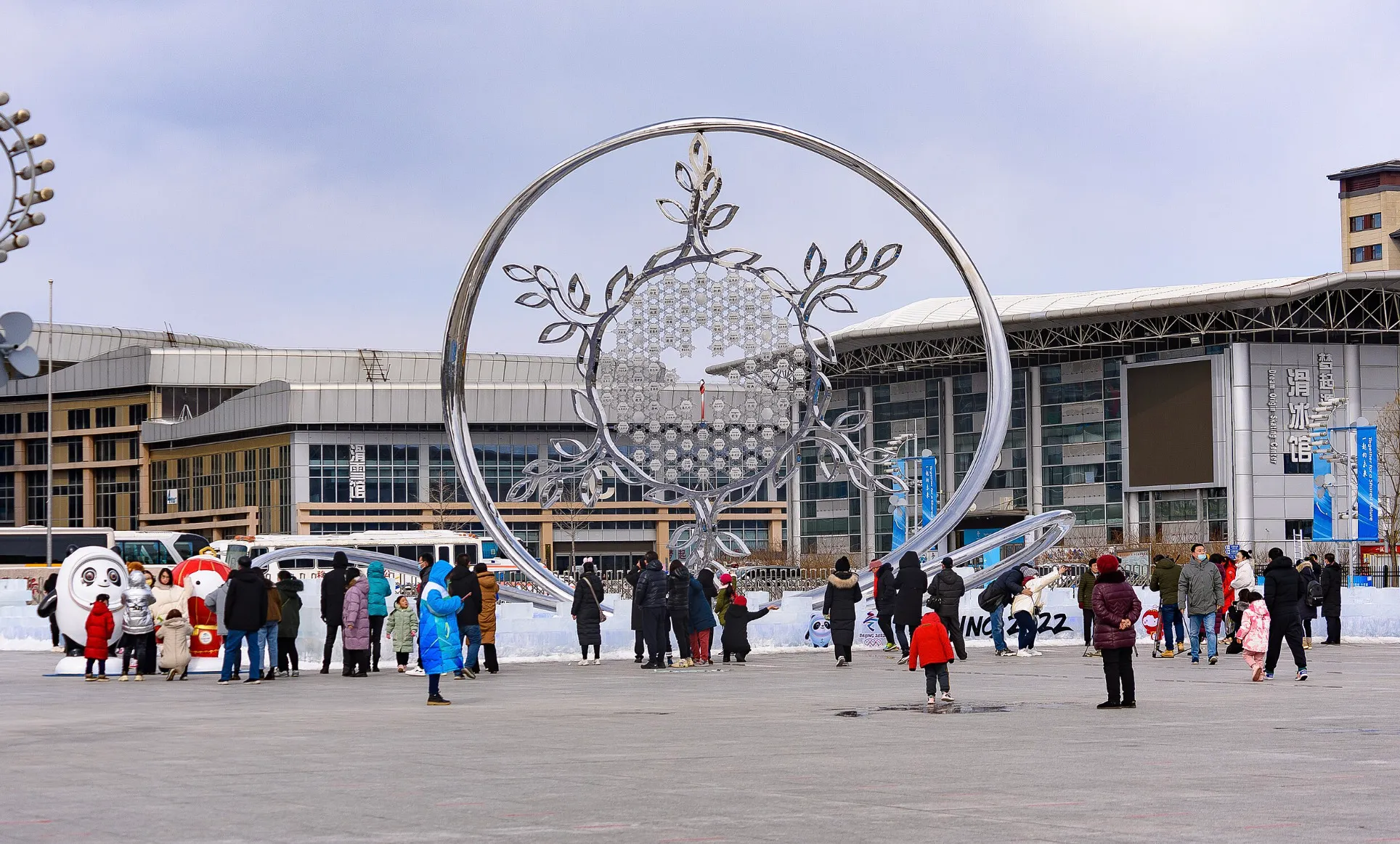 Visitors gather around a large metallic sculpture at an Olympic winter sports venue on a cold day.