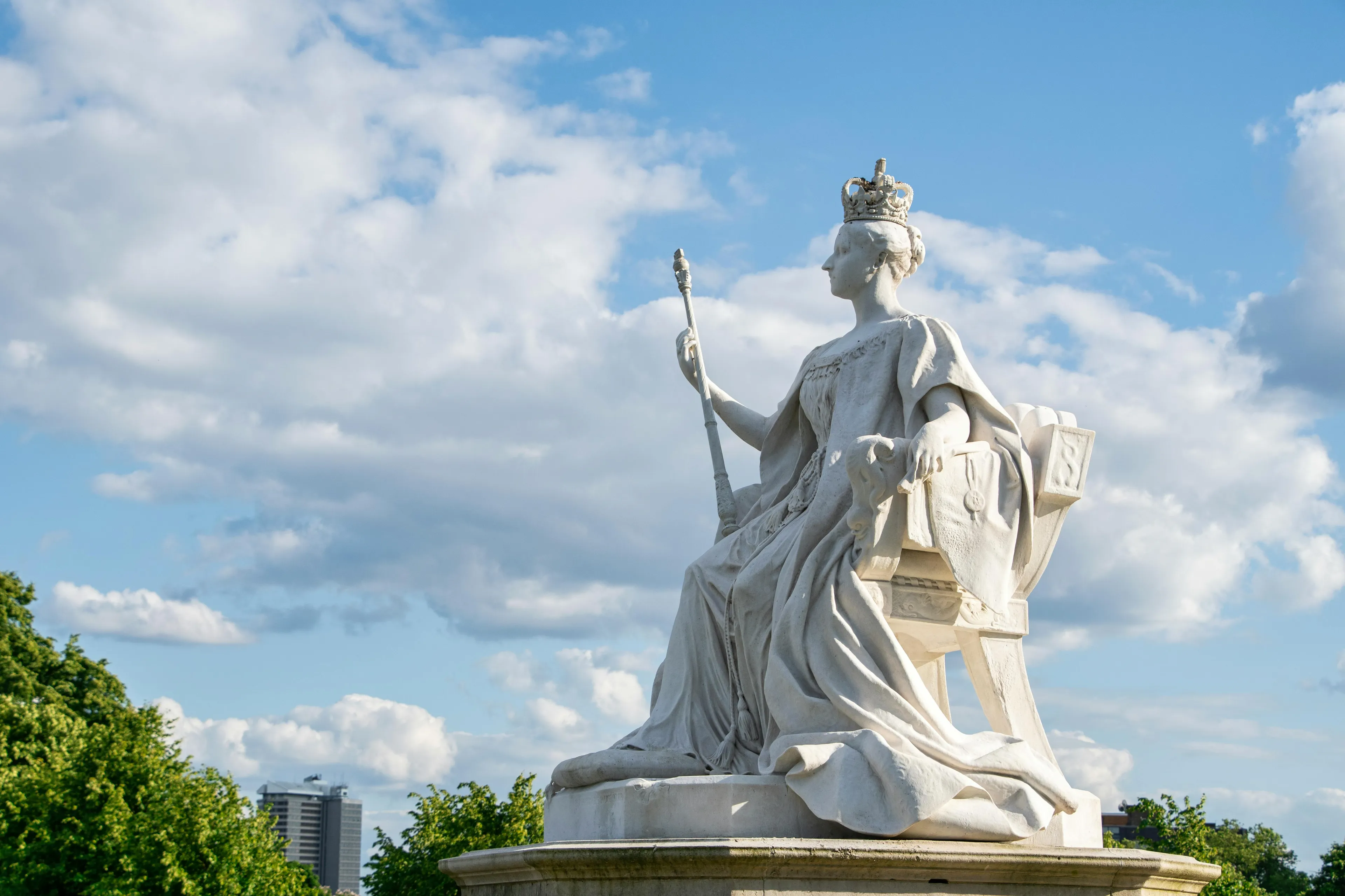 Statue of a queen regnant seated on a throne outdoors under a blue sky with clouds and greenery.