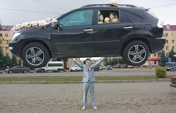 Novio en traje gris levantando un coche con decoración nupcial y novia dentro en una foto de boda inesperada.
