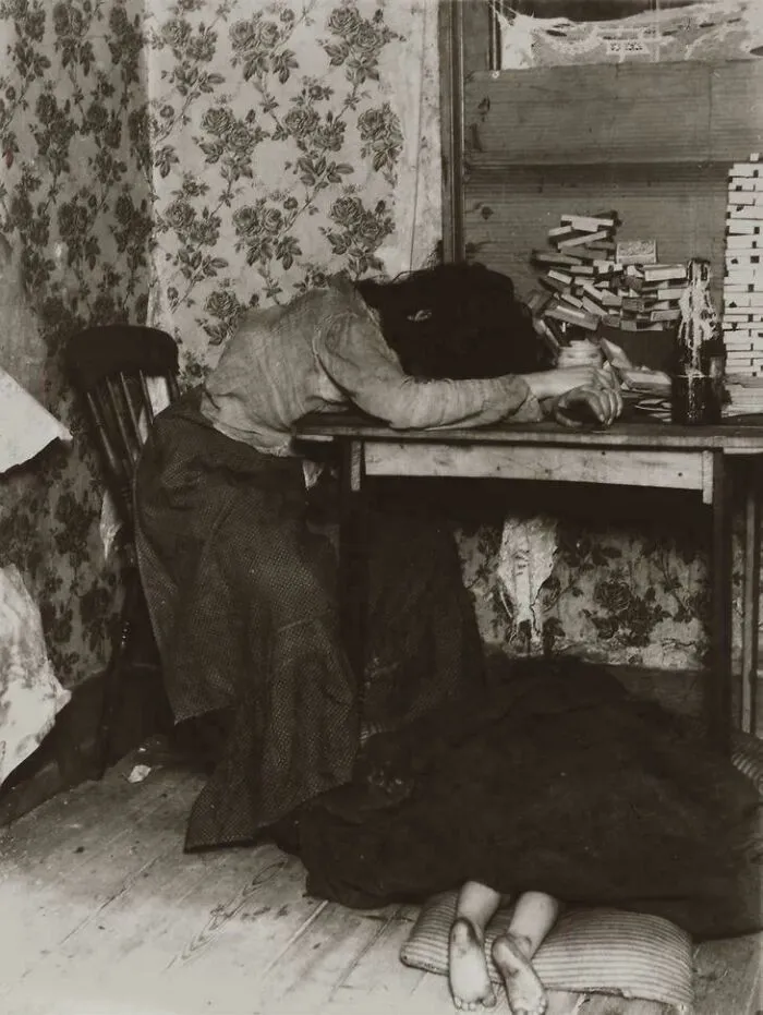 Woman resting head on table with cluttered workspace, highlighting harsh conditions in the Victorian era home environment.