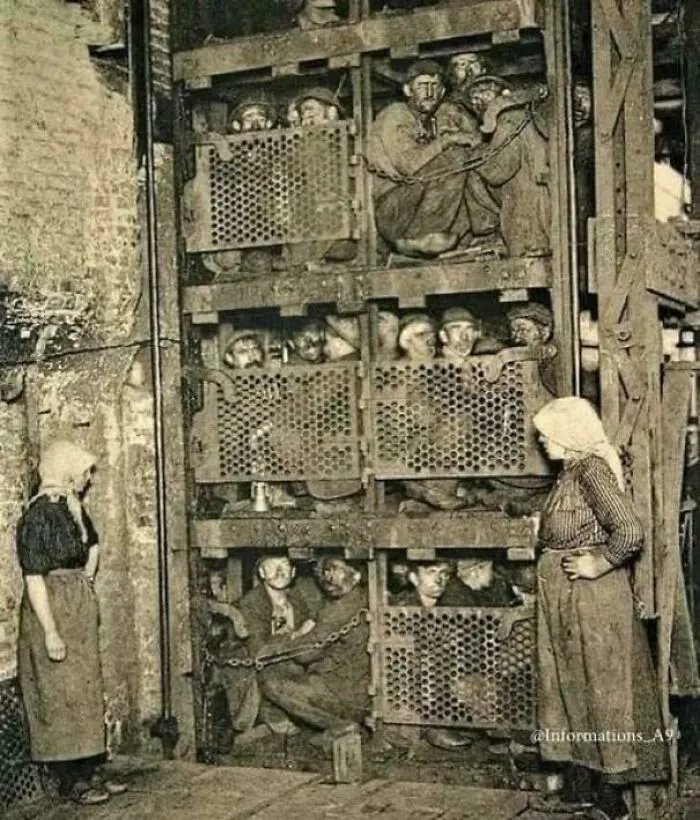 Victorian era coal miners crowded in a cage lift while women stand nearby in an industrial setting.
