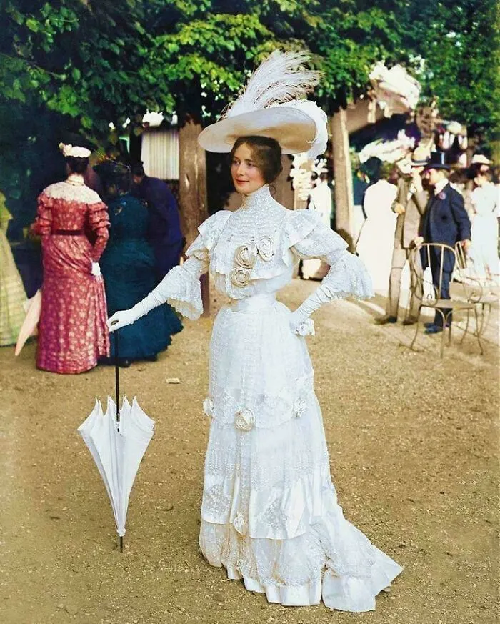 Victorian era woman in elegant white dress and large hat holding umbrella outdoors among other period-dressed people.