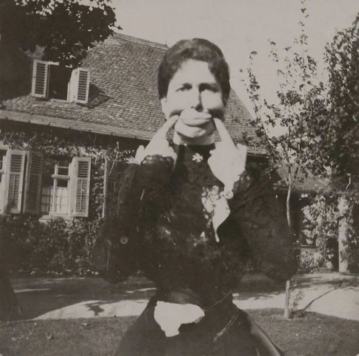 Victorian era woman making a funny face by pulling her mouth in a garden with a vintage house in the background.