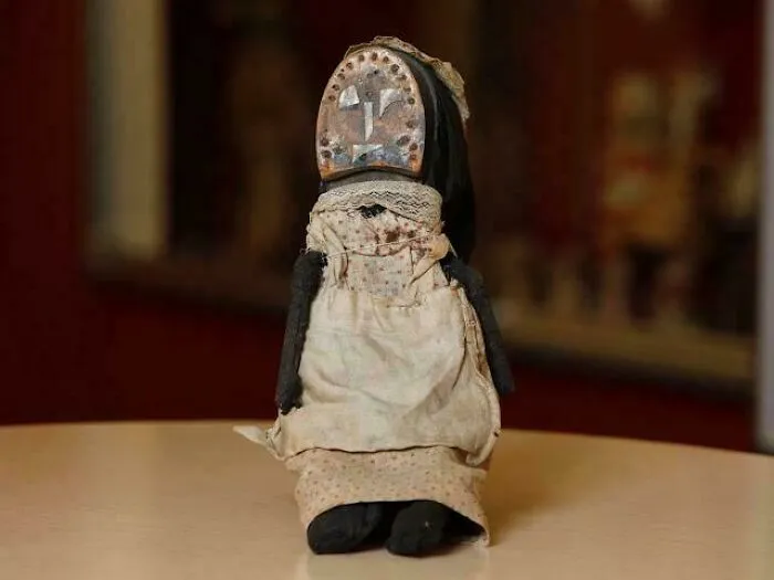 Worn Victorian era doll with fabric dress and painted face sitting on a table, showcasing vintage 19th century style.