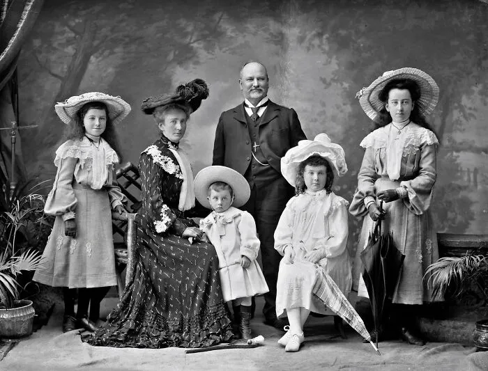 Victorian era family portrait with adults and children dressed in period clothing and large hats, holding umbrellas.