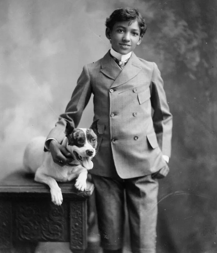 Victorian era boy in a double-breasted suit smiling next to a dog sitting on an ornately carved wooden table.