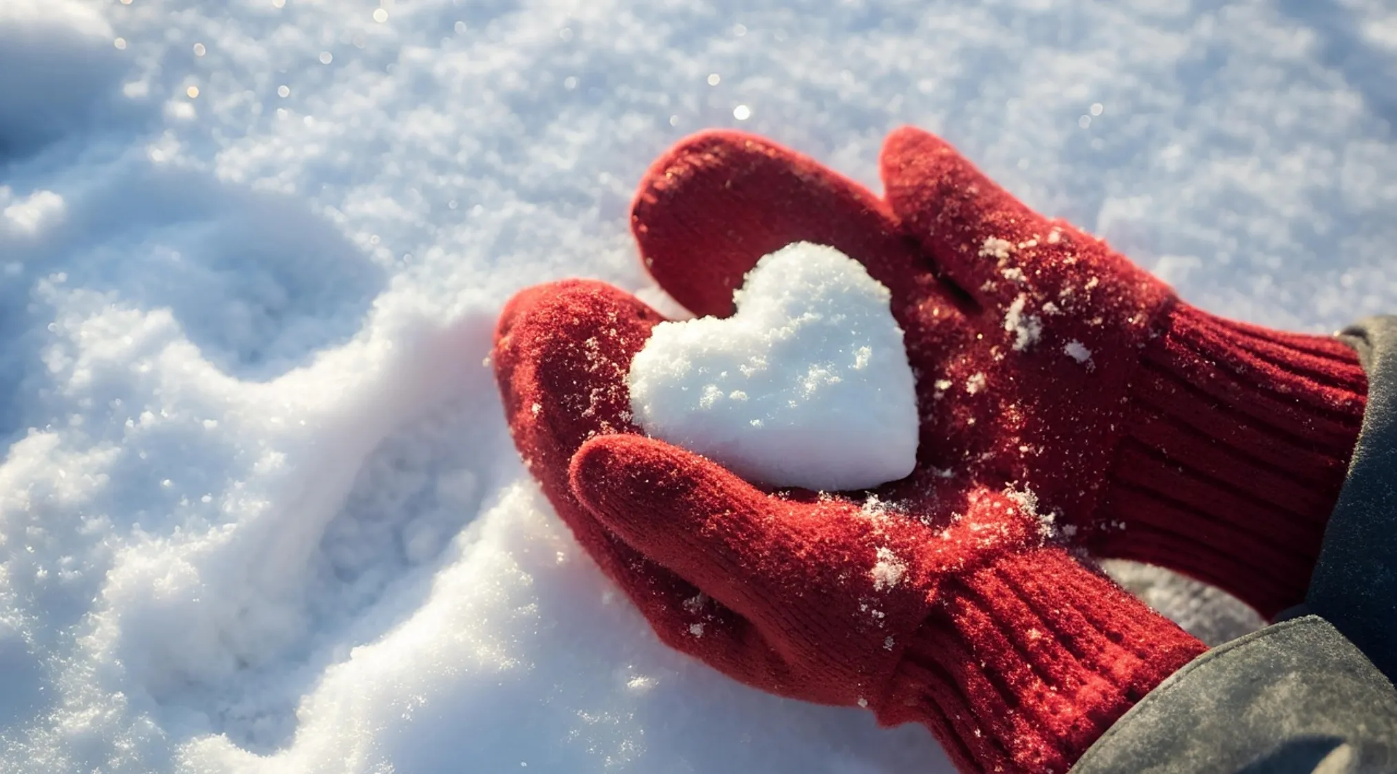 Hands in red gloves holding a snow heart shaped object in bright sunlight with surrounding snow background.