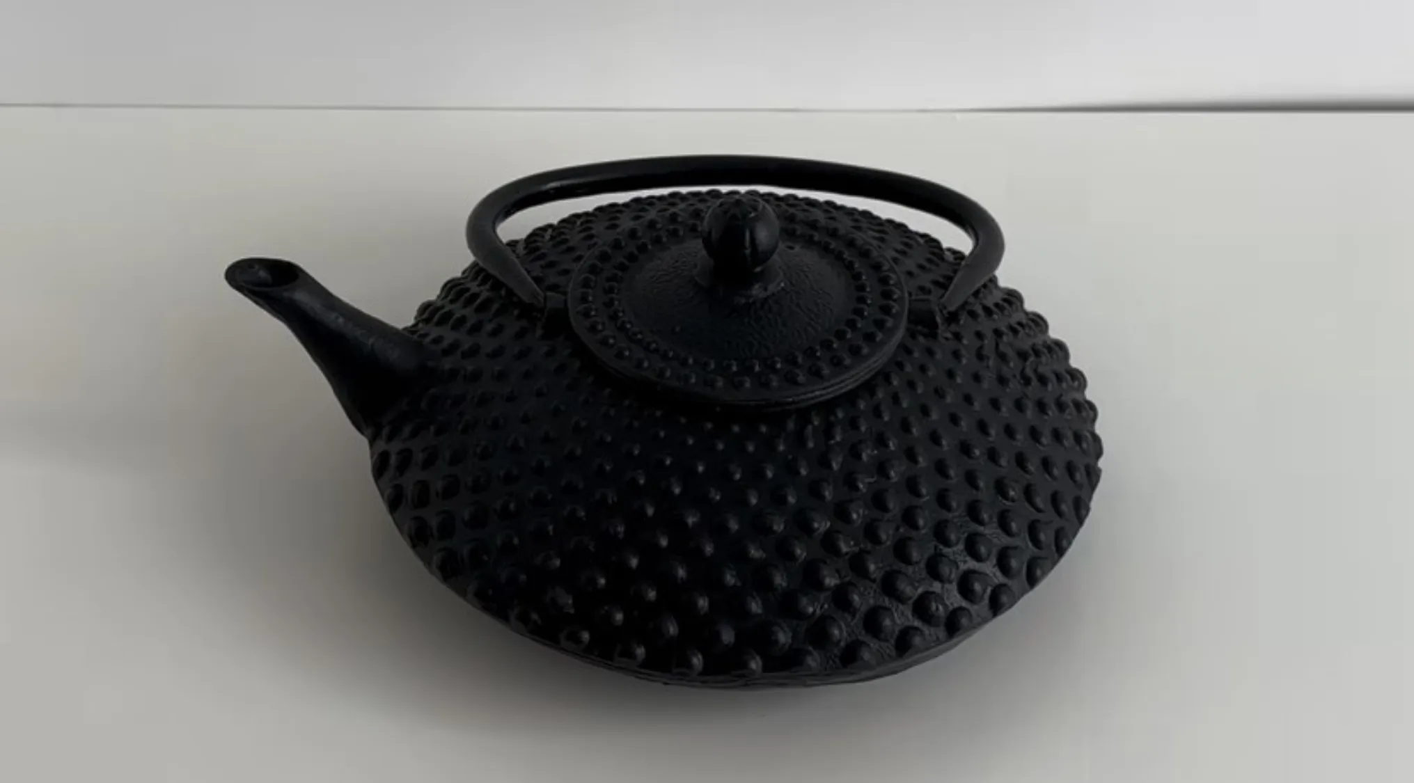 Black textured teapot with a handle and spout on a white surface, questioning if it is real or cake.
